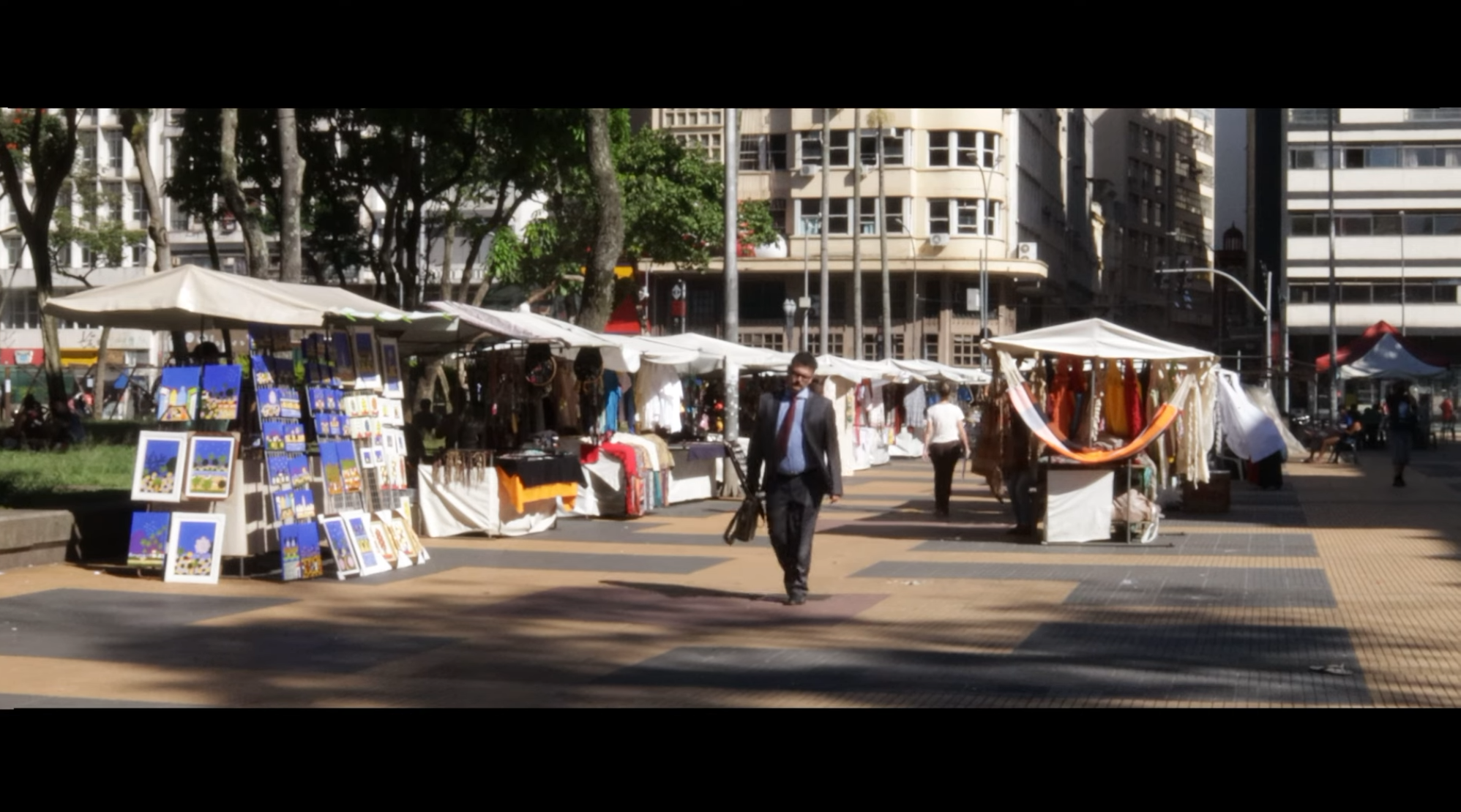 Cena de Afeto Shop, personagem caminhando entre barracas de uma praça em São Paulo